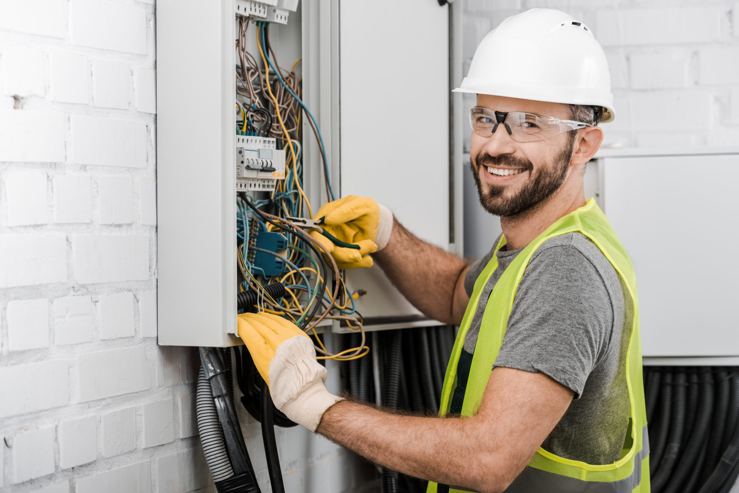An electrician wearing protective glasses and a hard hat, working on electric wiring. The image shows a professional electrician ensuring safety while performing electrical work