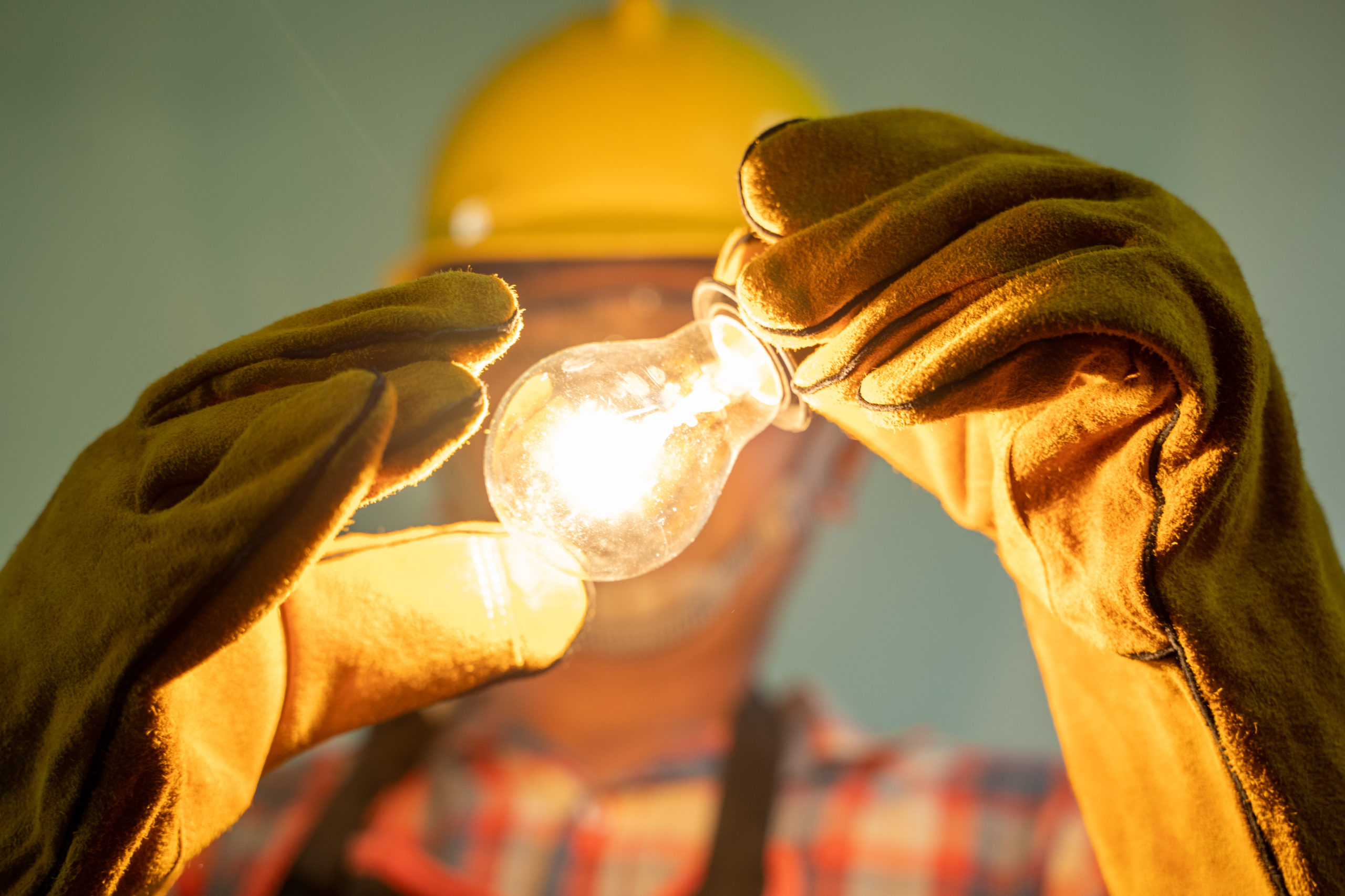 A man wearing hand gloves and a yellow hard hat, holding a light bulb that is switched on